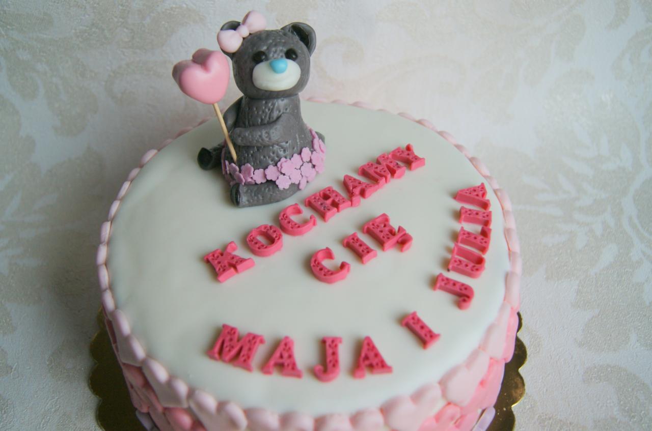 Tort urodzinowy z lukrowanym misiem trzymającym różowe serce, otoczony napisem 'Kocham Cię Maja i Julia' wykonanym z różowych literek.