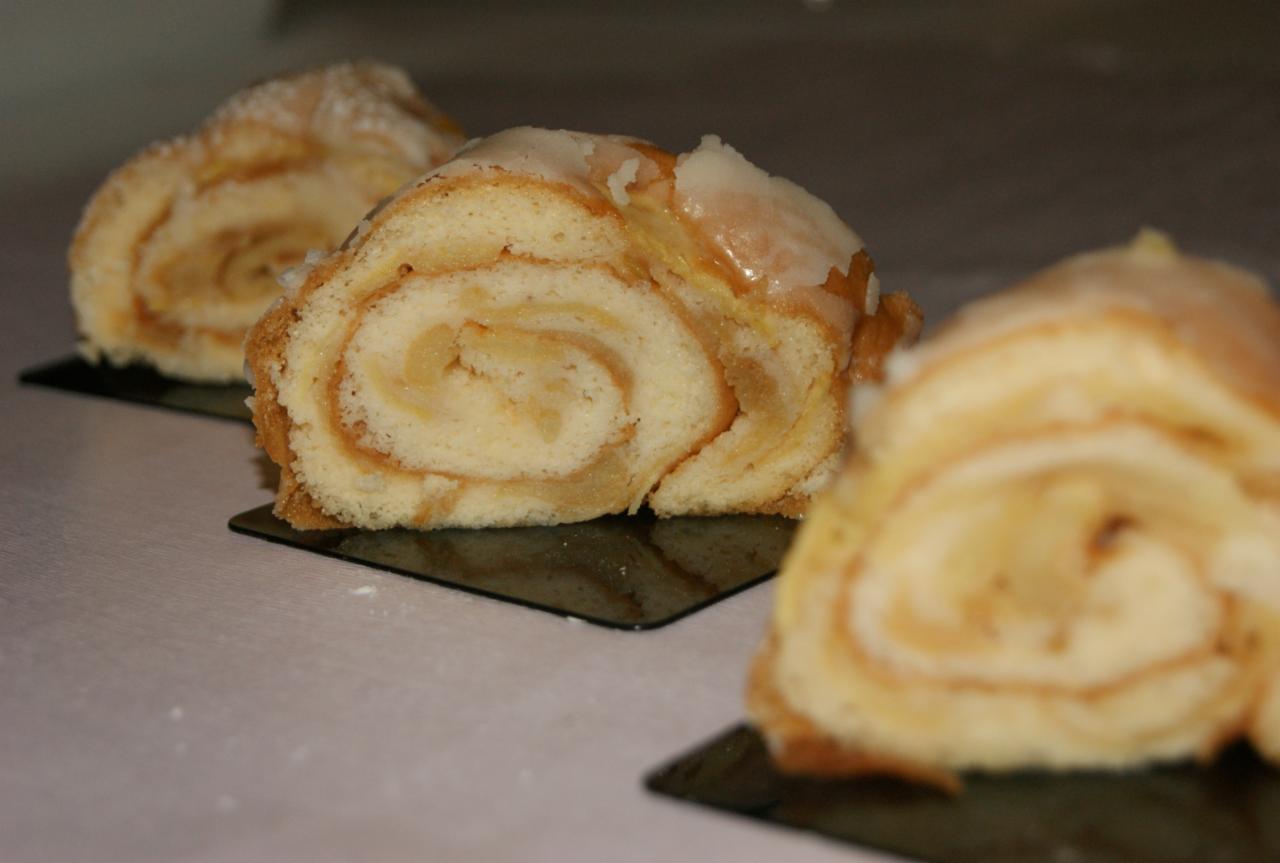 Trzy porcje rolad biszkoptowych z jasnym kremem i lukrem na czarnych, kwadratowych podstawkach, widoczne z bliska, w słabym oświetleniu.