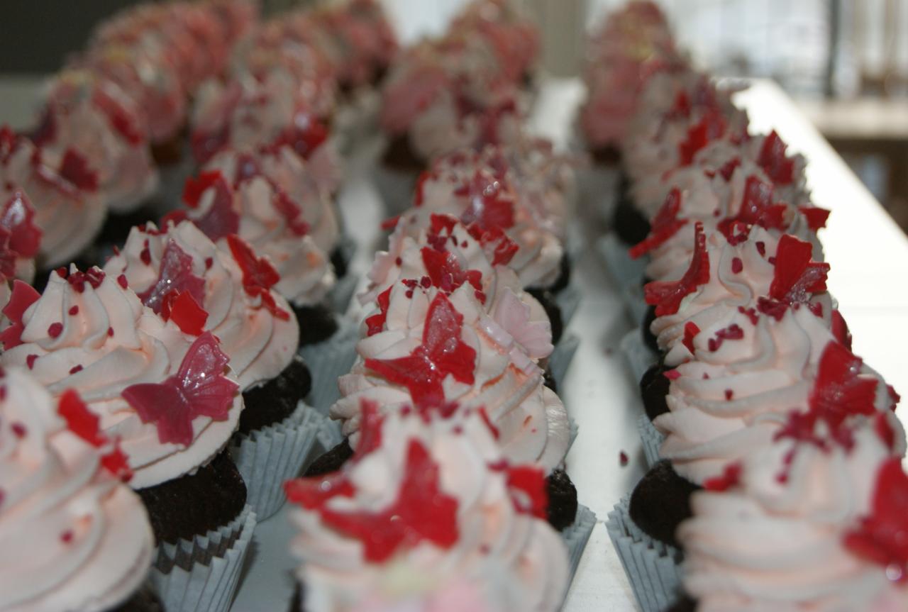 Rząd czekoladowych babeczek w papierowych foremkach, udekorowanych różowym kremem, czerwonymi posypkami i żelkowymi motylami.