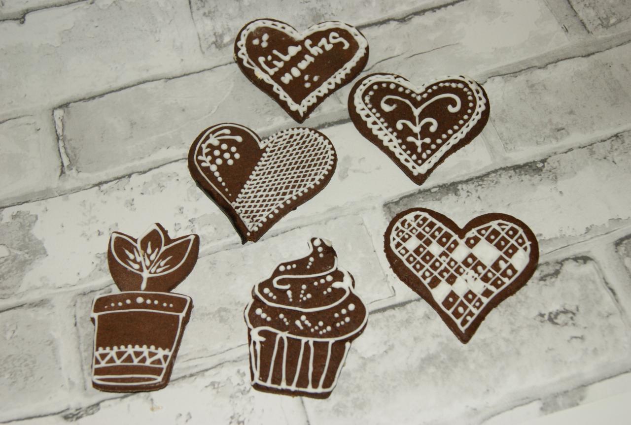 Ręcznie zdobione pierniczki w kształcie serc, babeczki i doniczki z kwiatkiem, leżące na tle imitującym cegły. Pierniczki posiadają białe lukrowe wzory, w tym napisy na jednym z serc.