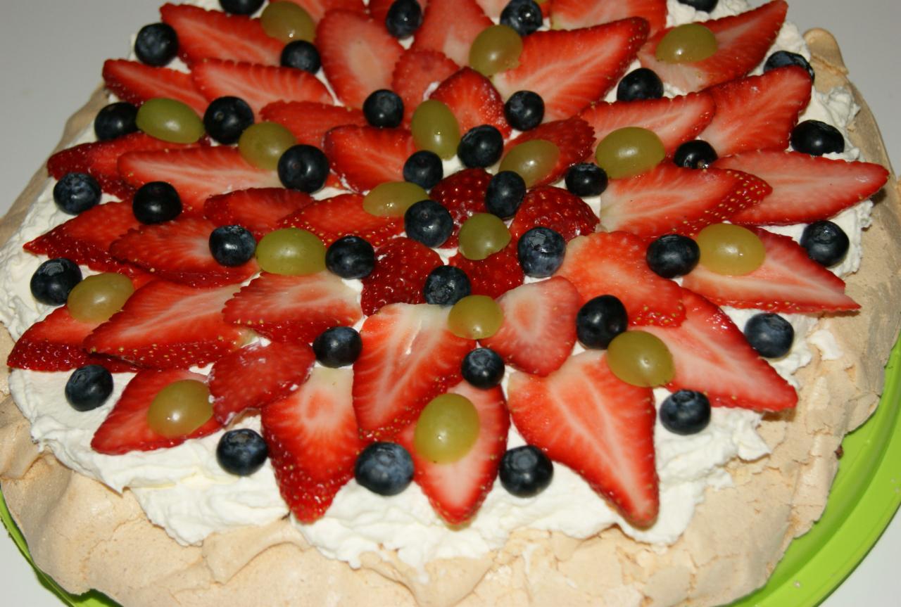 Tort bezowy udekorowany świeżymi truskawkami, borówkami i zielonymi winogronami, na białym kremie, z widoczną kruchą bezą.
