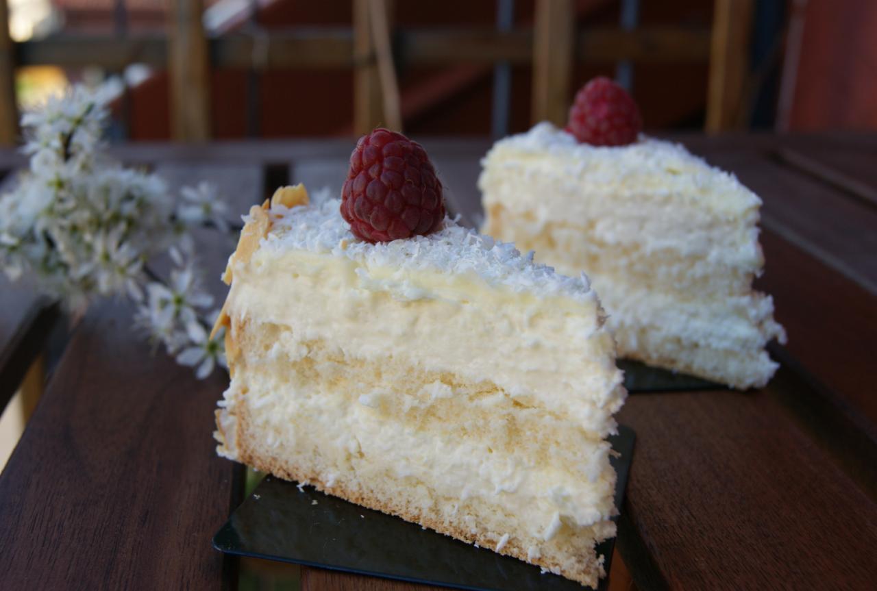 Dwa kawałki jasnego tortu z kremem i wiórkami kokosowymi, udekorowane malinami, na ciemnej podkładce, z gałązką kwitnącej wiśni w tle, na drewnianym stole.