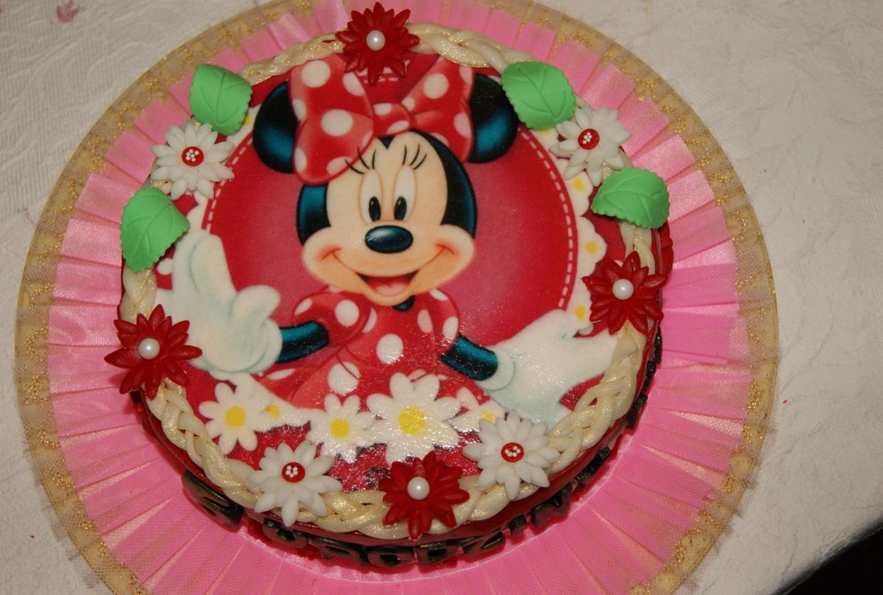 Tort urodzinowy z wizerunkiem Myszki Minnie, dekorowany kremowymi kwiatami, czerwonymi astrami z perłami i zielonymi listkami na różowym podkładzie z falbanką.