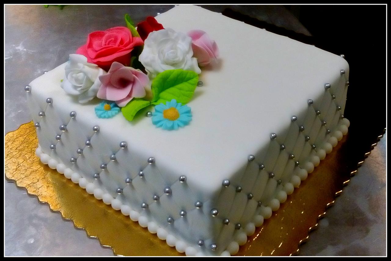 Kwadratowy tort z białym lukrem, ozdobiony srebrnymi perełkami w romby i bukietem cukrowych róż oraz błękitnych kwiatków, umieszczony na złotej podkładce.