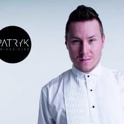 PERFORMA Patryk Gniedziejko - Portret mężczyzny w białej koszuli z czarnym logo 'Patryk Gnieździejko' na szarym tle, prezentujący profesjonalny wizerunek.