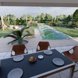 PERFORMA Patryk Gniedziejko - Nowoczesny ogród z zadaszonym basenem, drewnianym tarasem, huśtawką i altaną w kształcie kapsuły, widok z perspektywy stołu z nakryciem i laptopem.