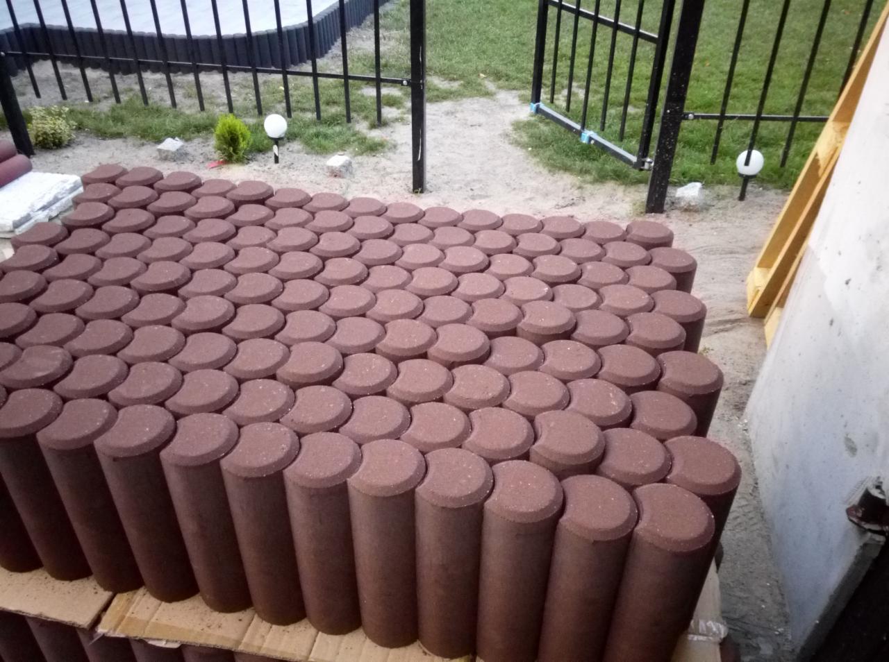 Czerwono-brązowe, cylindryczne elementy betonowe z półokrągłym wycięciem u góry, ułożone w rzędach na palecie, w tle czarne metalowe ogrodzenie i trawnik.