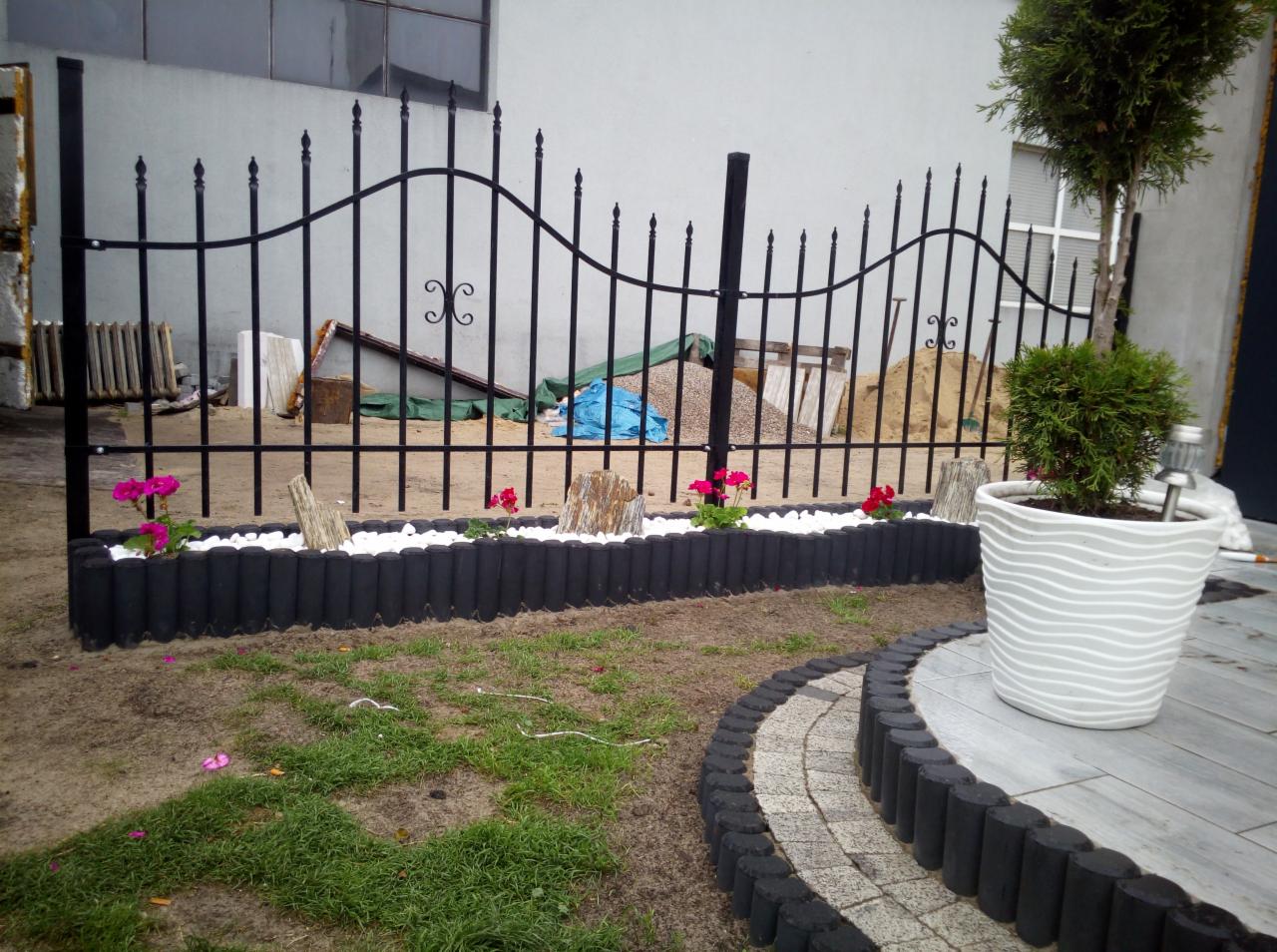 Czarne metalowe ogrodzenie z dekoracyjnym wykończeniem, rabata z białym kamieniem, kwiatami i drewnianymi elementami, obok ścieżka z obrzeżem i donica z rośliną.