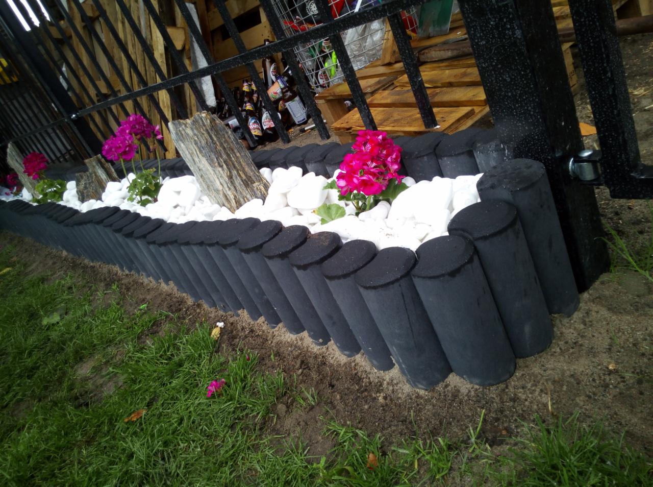 Niski płotek z betonowych palików w kolorze antracytowym, wypełniony białymi kamieniami i różowymi pelargoniami, widok z boku, w tle metalowa konstrukcja i paleta.