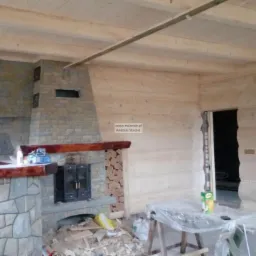 Wnętrze w trakcie wykończenia z kamiennym kominkiem, drewnianym barem i ścianami z jasnego drewna, widoczne narzędzia i materiały budowlane.