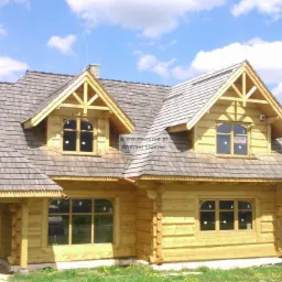 Surowy, jasny drewniany dom z bali z szarym dachem z gontów, widoczne okna z czarnymi ramami, w tle błękitne niebo z chmurami i zieleń.