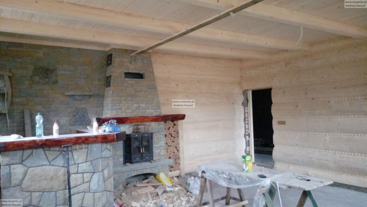 Wnętrze w trakcie wykończenia z kamiennym kominkiem, drewnianym barem i ścianami z jasnego drewna, widoczne narzędzia i materiały budowlane.