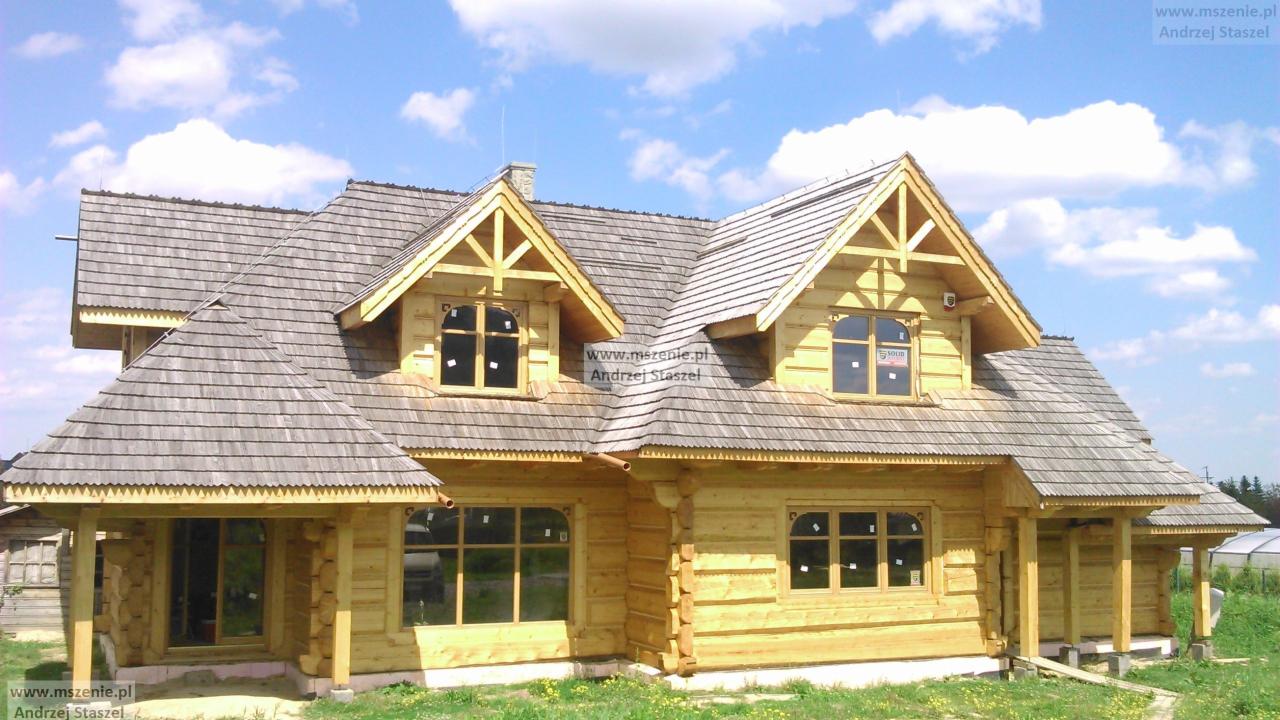 Surowy, jasny drewniany dom z bali z szarym dachem z gontów, widoczne okna z czarnymi ramami, w tle błękitne niebo z chmurami i zieleń.