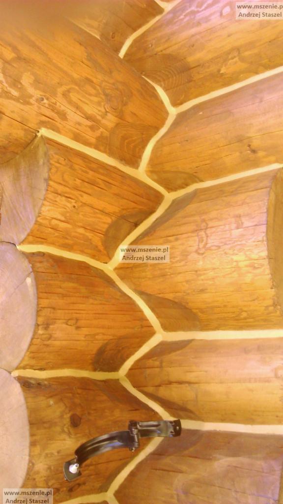 Wnętrze domu z belek, widoczne połączenia między belkami wypełnione jasną masą uszczelniającą oraz zamontowany metalowy element mocujący.