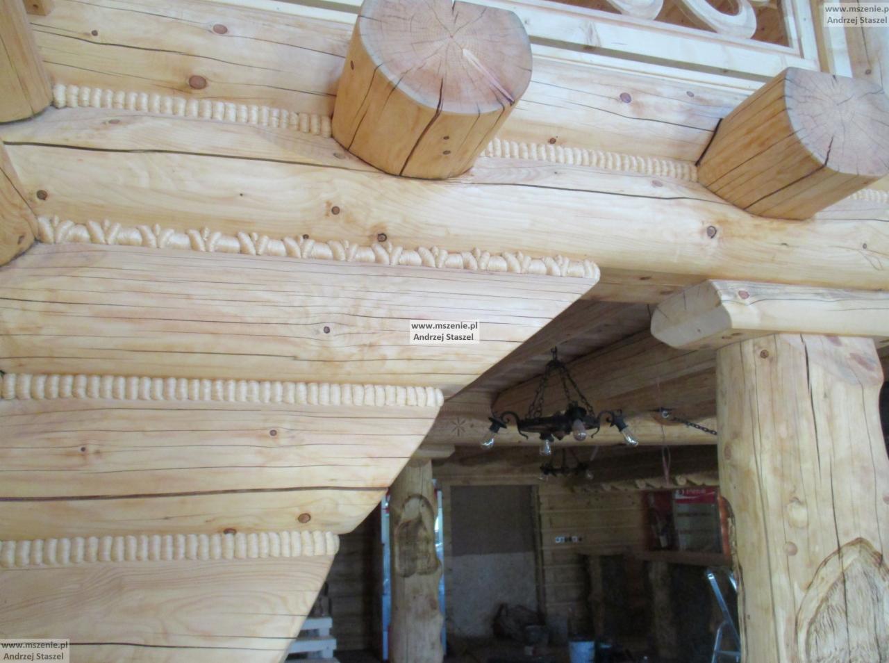 Wnętrze domu z bali z widoczną konstrukcją stropu, zdobieniami snycerskimi i żyrandolem w stylu retro.