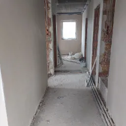 Korytarz w trakcie prac wykończeniowych w Rzeszowie: ściany częściowo pokryte tynkiem, widoczne instalacje elektryczne w ścianach, surowy betonowy sufit, podłoga w trakcie przygotowania.