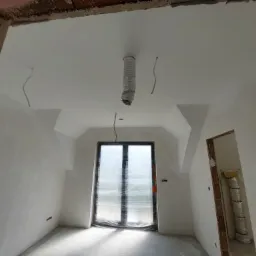 Pokój na poddaszu w trakcie prac wykończeniowych; widoczne gładkie, białe ściany, zabezpieczone okno folią, wystające kable i rura wentylacyjna zwisająca z sufitu.