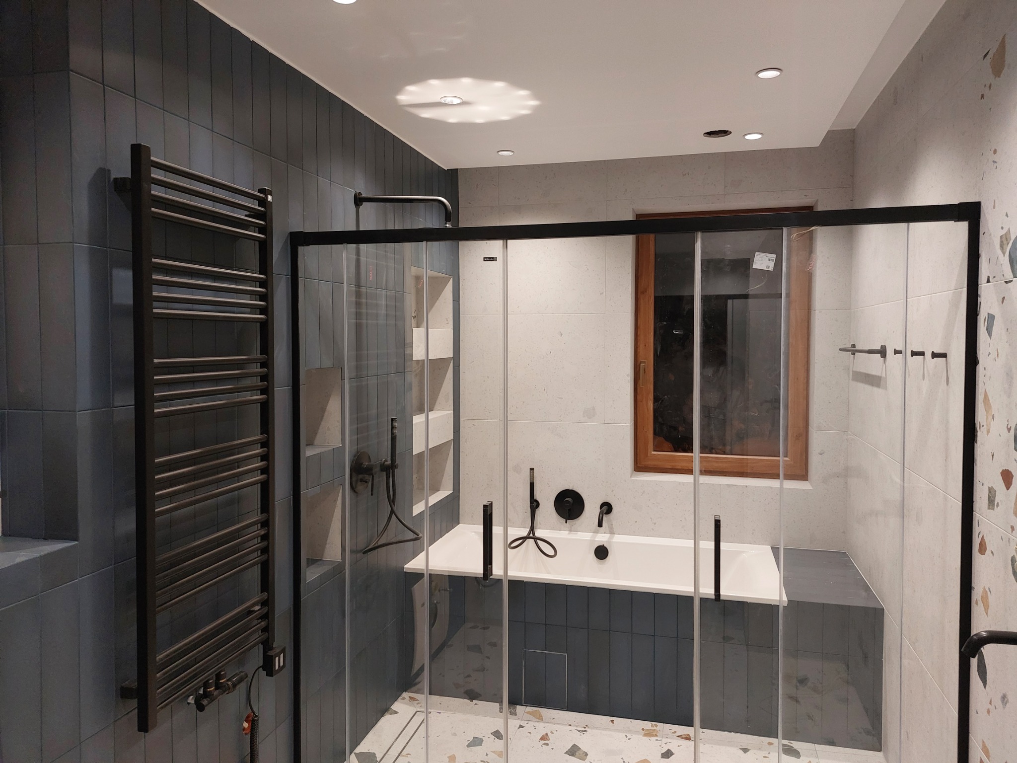 Nowoczesna łazienka z kabiną prysznicową typu walk-in, białą wanną obudowaną ciemnymi płytkami, grzejnikiem drabinkowym i podłogą w stylu lastryko.