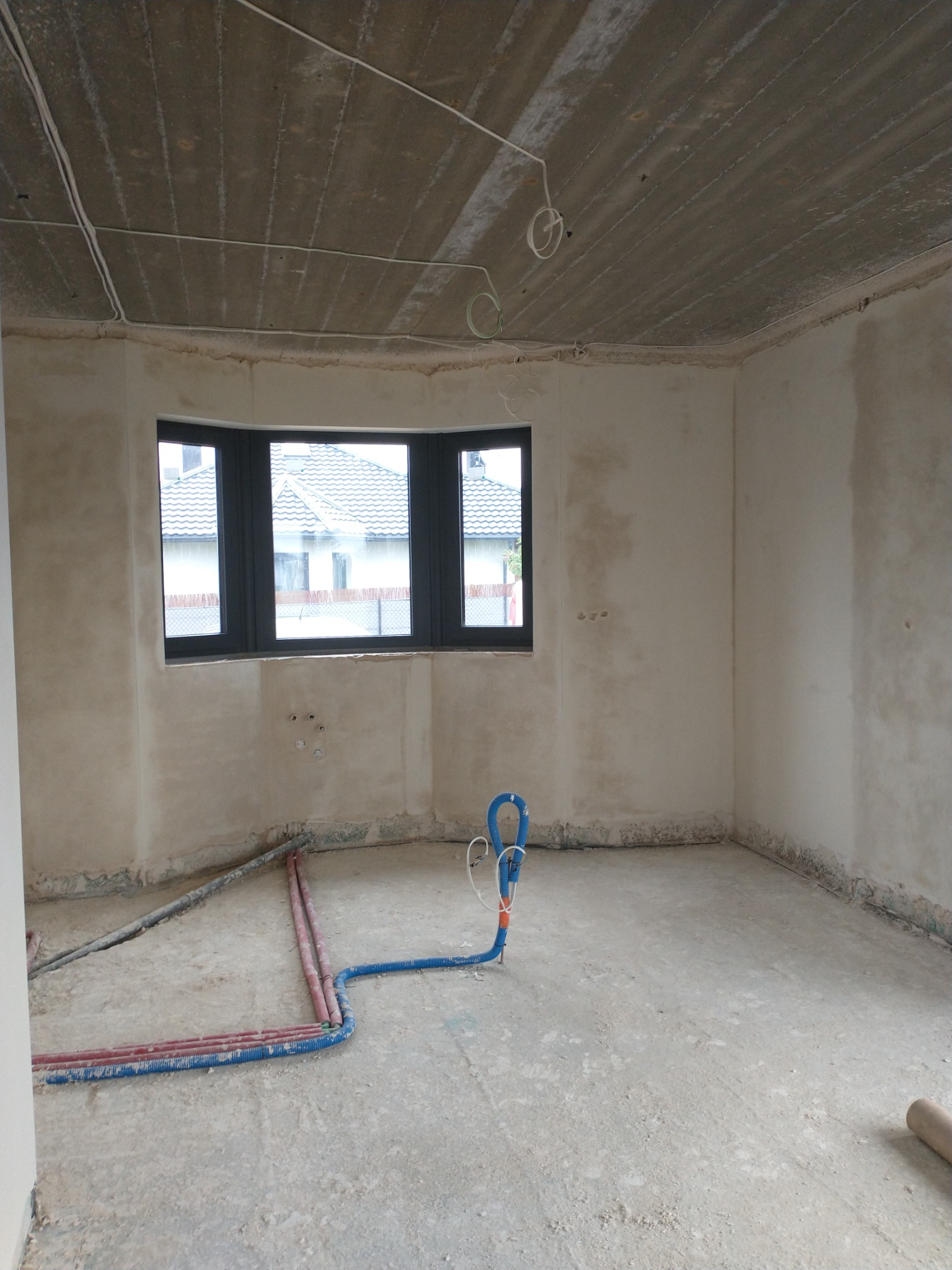 Surowy stan wnętrza z oknem wykuszowym, widocznymi instalacjami i nierówną powierzchnią ścian przygotowaną pod wykończenie.
