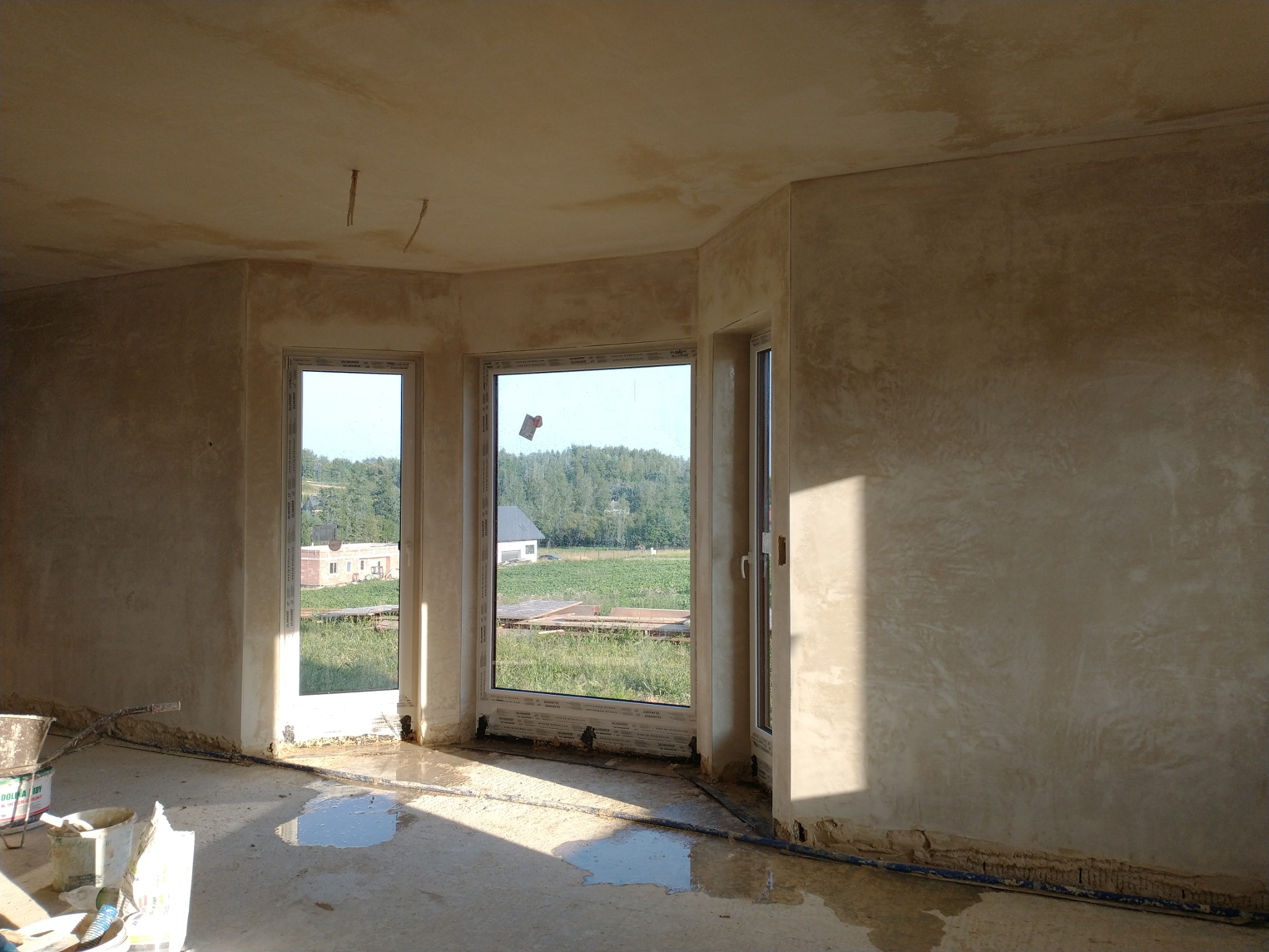 Wnętrze w trakcie prac wykończeniowych: ściany i sufit pokryte świeżo nałożoną, jasną warstwą, prawdopodobnie gładzią, z widocznymi śladami szpachlowania, plamy wody na podłodze, okna z widokiem...