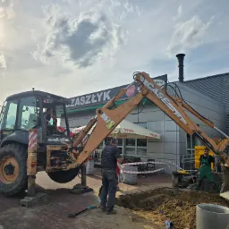 Koparko-ładowarka Case kopie d&oacute;ł na placu budowy, widoczni dwaj pracownicy w trakcie pracy. W tle budynek z szyldami reklamowymi i pochmurne niebo.