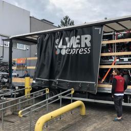 Silver Express Sp. z o.o. - Otwarta naczepa ciężarówki z logo Silver Express, załadowana motocyklami zabezpieczonymi pasami transportowymi, widoczny mężczyzna w czerwonej kamizelce i motocykl na zewnątrz, przed budynkiem.