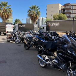 Silver Express Sp. z o.o. - Flota motocykli zaparkowana przed budynkiem, w tle widoczne samochody dostawcze firmy transportowej Silver Express i palmy.