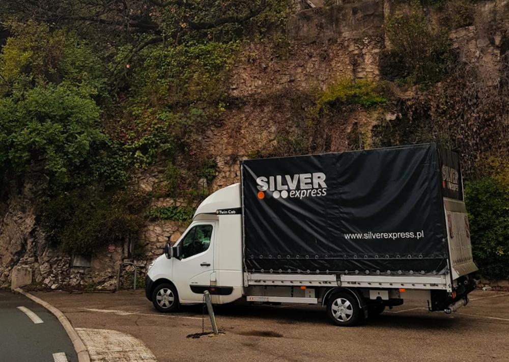 Biała furgonetka z czarną plandeką firmy Silver Express parkuje na tle kamiennego muru porośniętego zielenią.