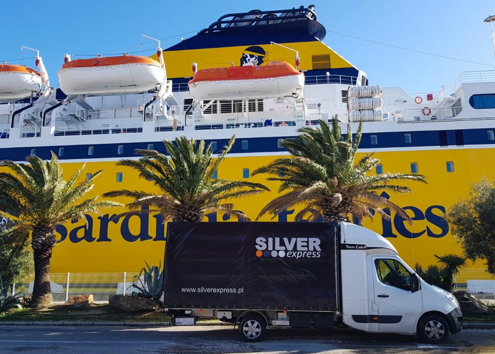 Biała furgonetka Silver Express z czarną plandeką na tle żółto-niebieskiego promu Sardinies i palm.