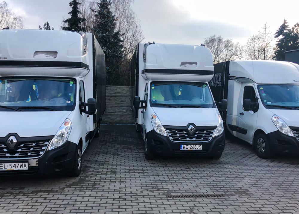 Trzy białe busy Renault Master z zabudową kontenerową, zaparkowane na brukowanym placu, z widocznymi tablicami rejestracyjnymi i logo firm transportowych.