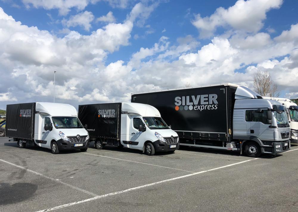 Flota pojazdów firmy Silver Express: dwa białe busy Renault Master z czarnymi plandekami i duża ciężarówka MAN z czarną naczepą, zaparkowane na parkingu pod błękitnym niebem z chmurami.