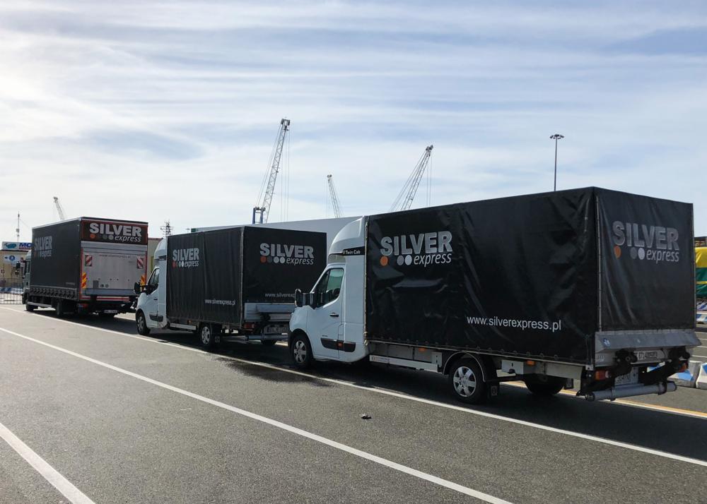Trzy białe samochody dostawcze firmy Silver Express z czarnymi plandekami na naczepach, zaparkowane na betonowej nawierzchni portu, z dźwigami w tle.