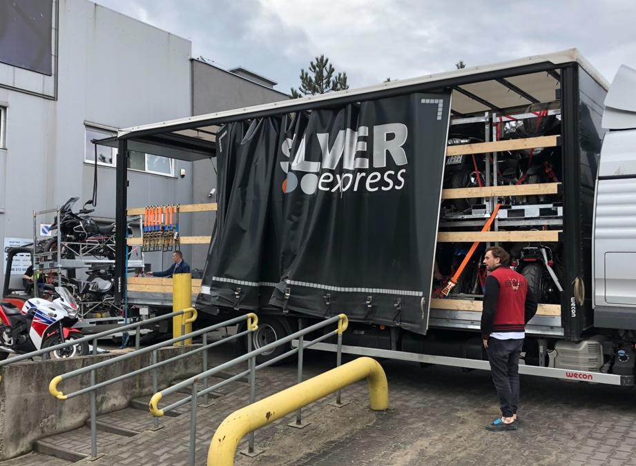 Otwarta naczepa ciężarówki z logo Silver Express, załadowana motocyklami zabezpieczonymi pasami transportowymi, widoczny mężczyzna w czerwonej kamizelce i motocykl na zewnątrz, przed budynkiem.