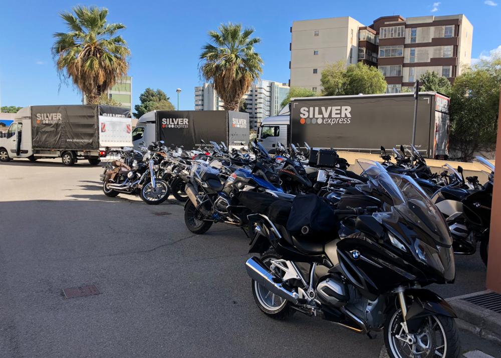 Flota motocykli zaparkowana przed budynkiem, w tle widoczne samochody dostawcze firmy transportowej Silver Express i palmy.