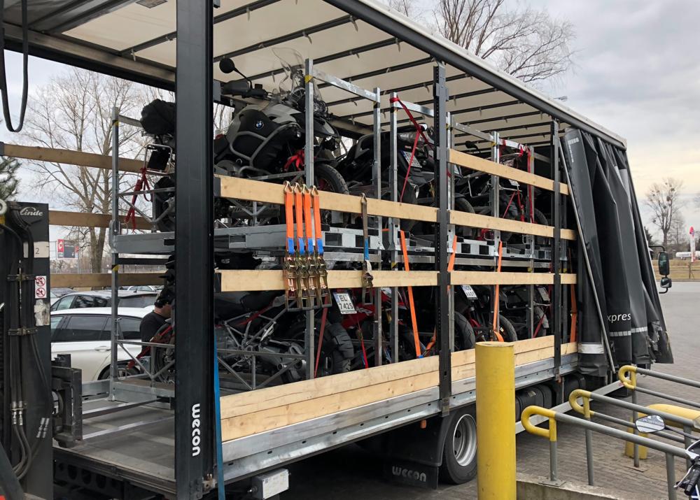 Ciężarówka z otwartą naczepą przewozi kilka motocykli BMW GS zabezpieczonych pasami transportowymi, widoczne drewniane zabezpieczenia i metalowa konstrukcja naczepy.