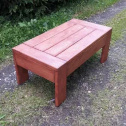 Prostokątny, drewniany stolik kawowy w odcieniach brązu, z blatem składającym się z desek, umieszczony na zewnątrz na ścieżce z trawiastym poboczem i zielonymi krzewami w tle.