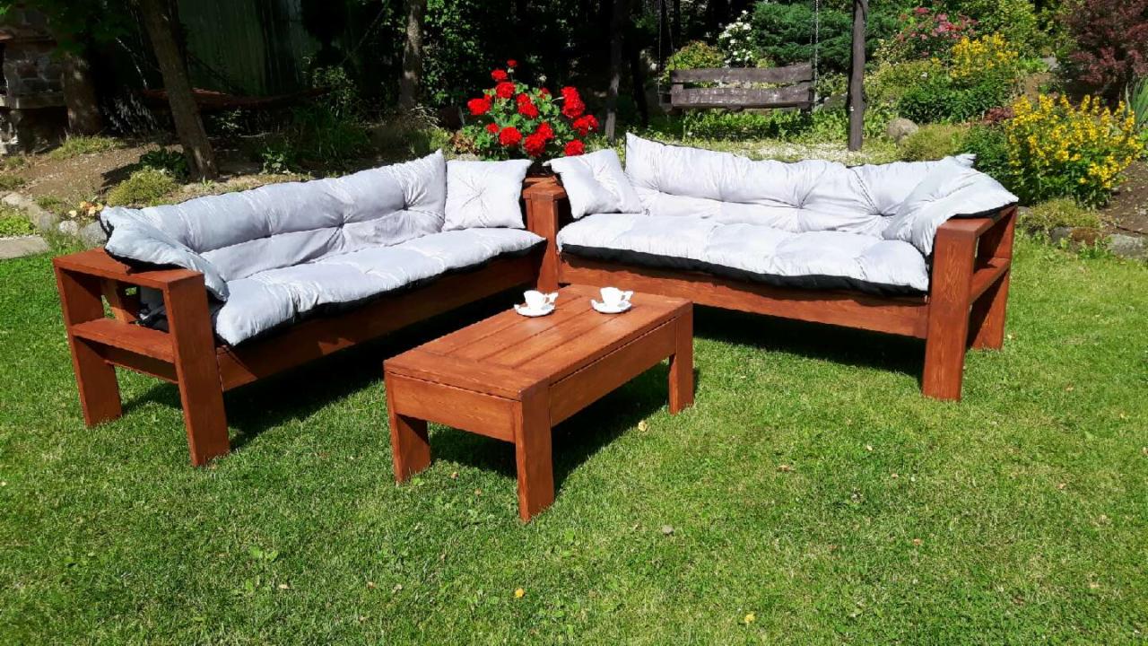 Dwa drewniane fotele ogrodowe z szarymi poduszkami i stolik kawowy z dwoma filiżankami na trawniku, w tle rabata z czerwonymi różami i huśtawka.