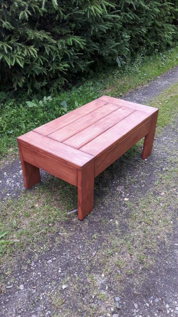 Prostokątny, drewniany stolik kawowy w odcieniach brązu, z blatem składającym się z desek, umieszczony na zewnątrz na ścieżce z trawiastym poboczem i zielonymi krzewami w tle.