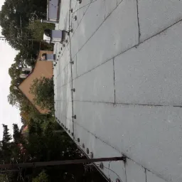 Szary, płaski dach pokryty papą termozgrzewalną z widocznymi mocowaniami i elementami wentylacyjnymi, w tle drzewa i budynek.