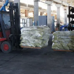 Czerwony wózek widłowy Linde transportuje paletę z workami zapakowanymi w folię stretch w hali magazynowej ze słupami i konstrukcją stalową