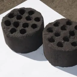 Dwa cylindryczne brykiety z otworami, ułożone na białym papierze na szarym, betonowym podłożu.