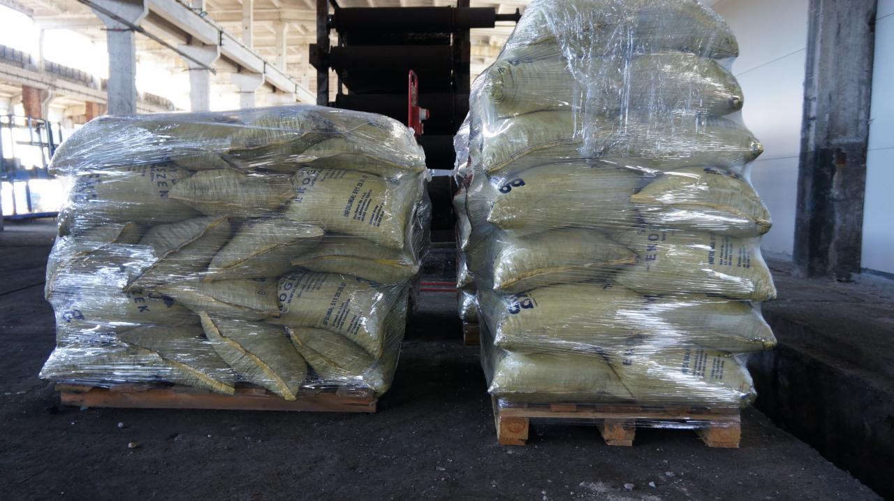 Dwa stosy worków z opałem, owinięte folią stretch, ułożone na drewnianych paletach w hali magazynowej z betonowymi filarami i elementami konstrukcyjnymi pod sufitem.