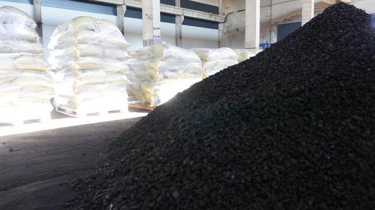 Stos czarnego węgla kamiennego w magazynie, worki z towarem owinięte folią na paletach w tle, betonowe filary konstrukcyjne hali.