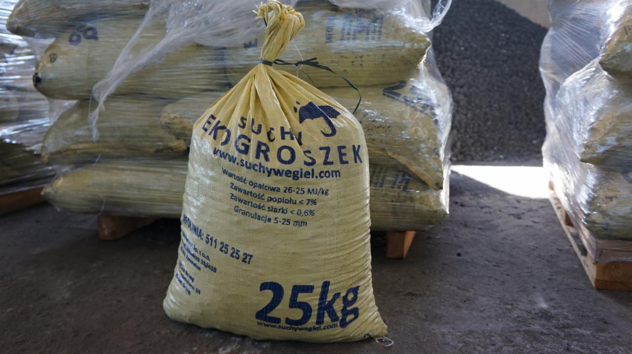 Żółty, foliowy worek 25 kg z ekogroszkiem 'Suchy Groszek' na tle palety z workami, widoczne dane techniczne i kontaktowe producenta.