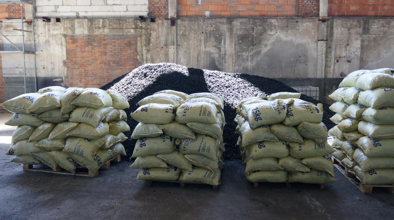 Stosy worków z opałem na paletach, za nimi góra czarnego materiału, w tle ceglana ściana magazynu.