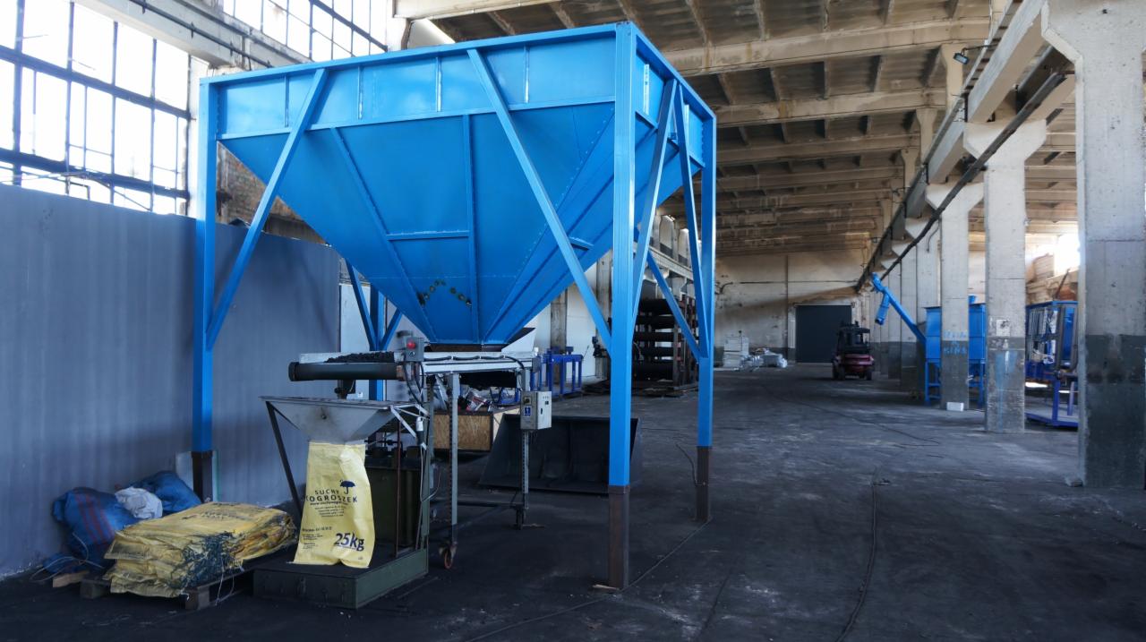 Niebieski zasobnik na węgiel z workami 25kg w hali produkcyjnej z betonowymi kolumnami i dużymi oknami.