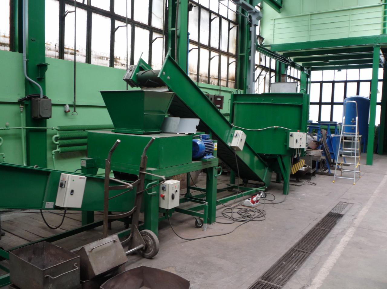 Widok na linię produkcyjną z zielonymi elementami, w tle okna fabryczne i metalowa drabina oparta o niebieski zbiornik.