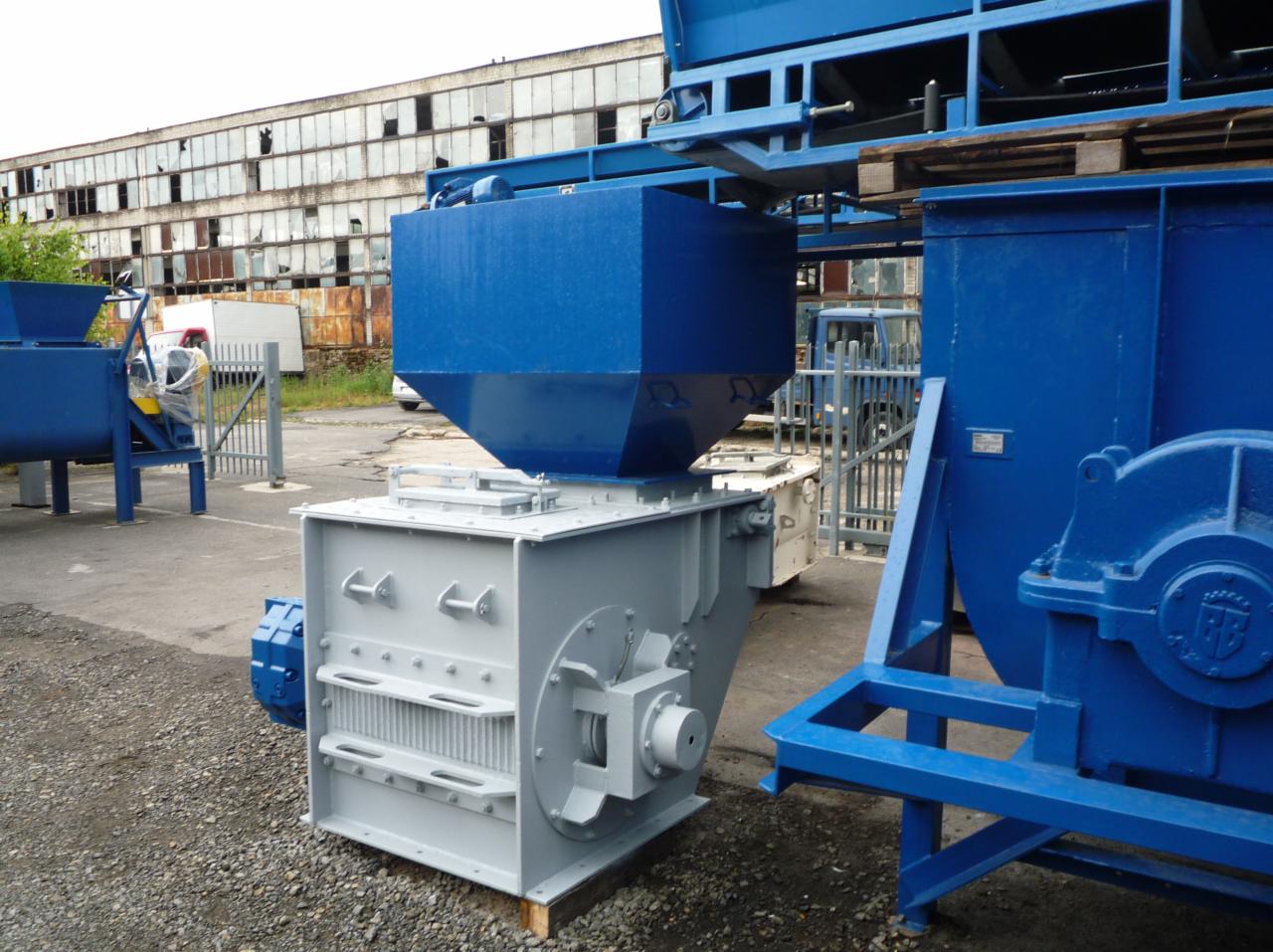 Szaro-niebieska maszyna do kruszenia węgla na tle zniszczonego budynku przemysłowego i zaparkowanych samochodów dostawczych.