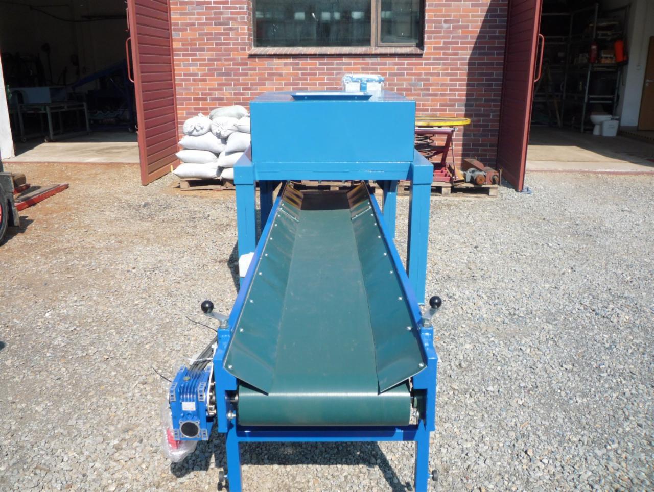 Niebieska maszyna z taśmą transportującą na zewnątrz, obok sterta worków i otwarte wrota magazynu z ceglaną ścianą w tle.