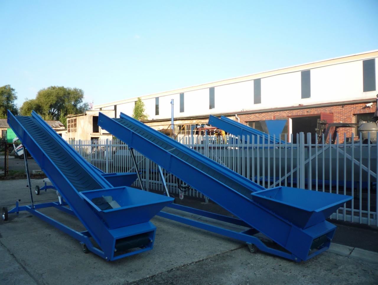 Dwa mobilne podajniki taśmowe w kolorze niebieskim, na zewnątrz, z niskim kątem ujęcia, ustawione na betonowej powierzchni przed budynkiem.